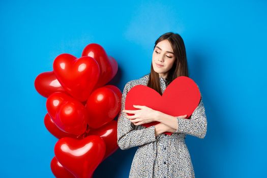 Valentines day. Romantic pretty woman in dress hugging big red heart cutout and looking dreamy, thinking of love, standing on blue background.