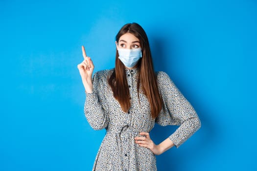 People, covid and quarantine concept. Excited cute female in medical mask, pointing and looking at logo with happy amazed face, standing on blue background.