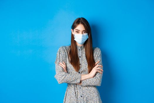Covid-19, pandemic lifestyle concept. Serious young woman with long natural hair, wearing medical mask, cross arms on chest, protect herself from virus, blue background.