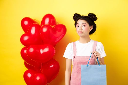 Happy Valentines day. Stylish asian single girl buying herself gift, holding shopping bag and looking unbothered at camera, standing over yellow background.
