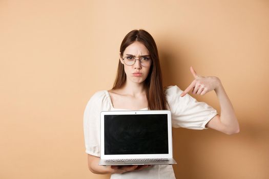 Angry frowning woman in glasses pointing at laptop screen, complaining and look disappointed, standing on beige background.