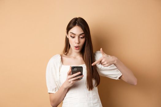 Exciting news on phone. Surprised cute woman reading online promo on screen, pointing at smartphone and say wow amazed, standing on beige background.