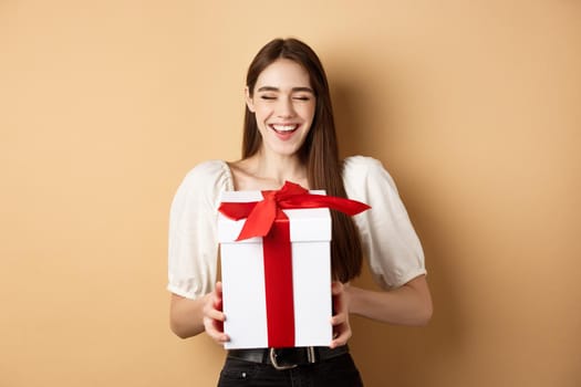 Happy smiling woman laughing, holding gift box from lover, enjoying romantic date on Valentines day, standing on beige background.