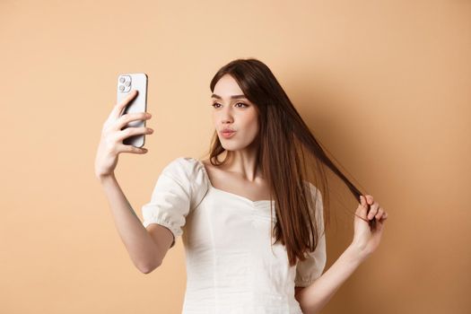 Stylish young woman playing with hair and taking selfie on smartphone, make photo for social media, standing on beige background.