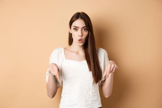 Wow amazing. Impressed young woman look astonished by product, pointing fingers down at logo, standing on beige background.