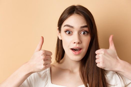 Very cool. Excited girl showing thumbs up and gasping amazed, praising good job, compliment something awesome, standing on beige background.