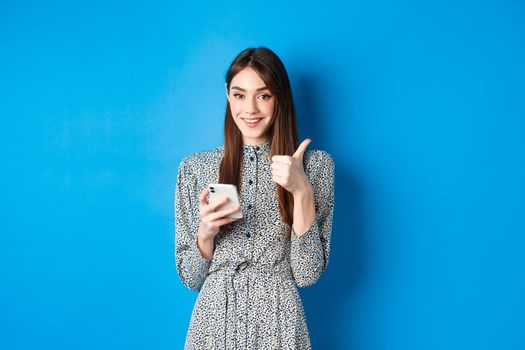 Happy smiling girl in stylish dress, showing thumb up in approval, using smartphone app and recommend it, standing against blue background.