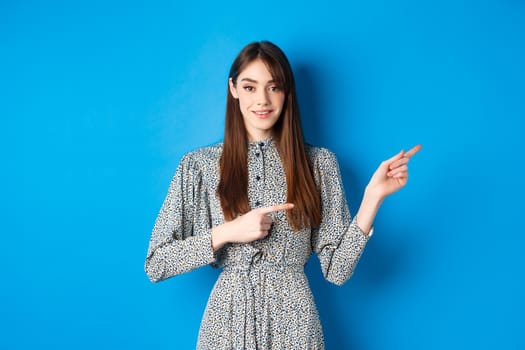 Young attractive woman in dress, with long natural hair, pointing fingers right at logo, smiling and inviting check out promotion, standing on blue background.