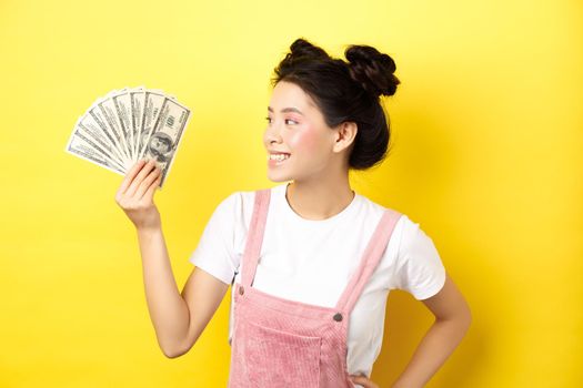Shopping. Rich and stylish asian female model showing money, looking at dollar bills with pleased smile, standing on yellow background.