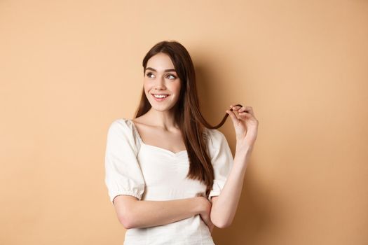 Romantic pensive girl thinking, looking left at empty space and playing with hair strand, smiling dreamy, standing in white dress on beige background.