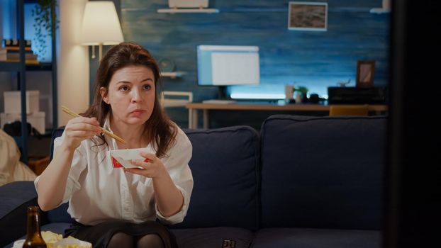 Person eating asian noodles with chopsticks on couch sitting in living room. Caucasian woman with chinese food box from delivery package enjoying meal while watching movie on TV screen