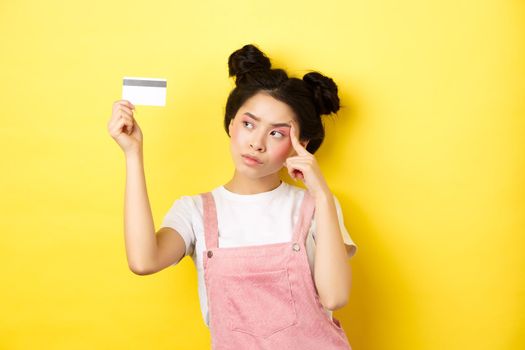 Shopping. Thoughtful trendy girl with summer makeup, looking pensive at plastic credit card, standing on yellow background.