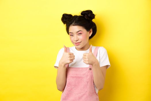 Cute teen asian girl with beauty makeup, showing thumbs up and smiling, praising and motivating you, making compliment, standing pleased on yellow background.