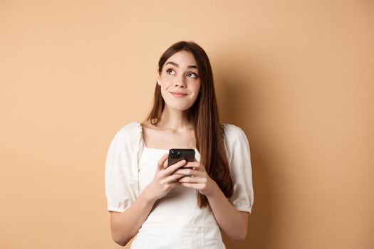 Romantic girl thinking with phone, looking aside and smiling dreamy, using dating app on smartphone, standing on beige background.