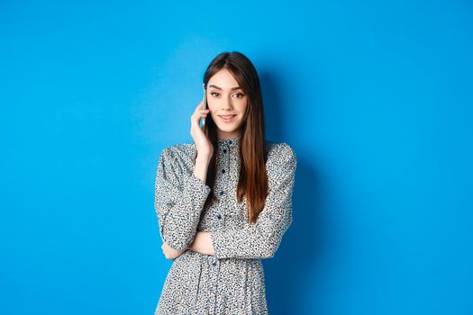 Stylish young woman talking on phone, making takeout order via smartphone, standing in dress against blue background.