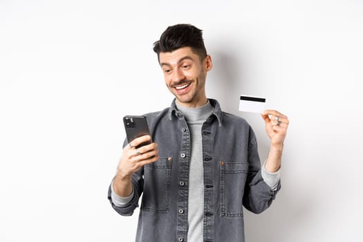 Online shopping concept. Handsome man buying in internet, showing plastic credit card and look at smartphone, standing against white background.