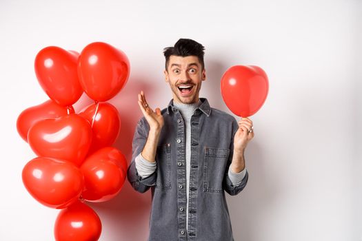 Valentines day concept. Cheerful young man celebrating love holiday, standing near red heart balloons and looking surprised, scream of joy, white background.