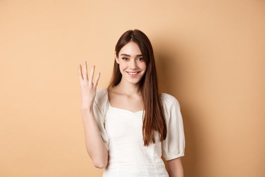 Attractive young woman show fingers number four, smiling and looking confident, standing on beige background.
