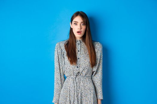 Shocked cute girl in dress looking at camera with opened mouth, gasping startled, see something shocking, standing on blue background.