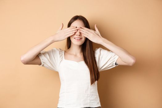 Happy romantic girl covering eyes and waiting for surprise, smiling while anticipating gift, standing on beige background.