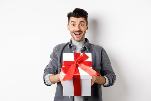Surprised young man smiling excited, holding big gift box on valentines day holiday, receive surprise present, standing amazed on white background.