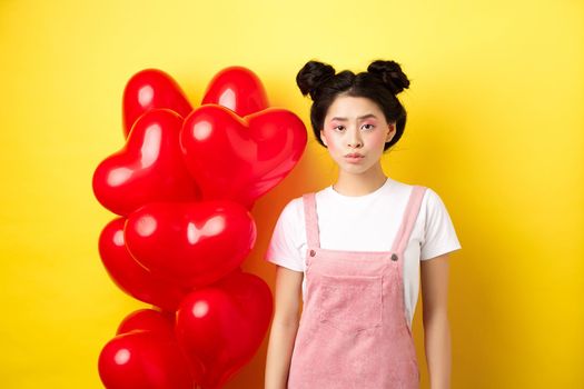 Valentines day concept. Sad and gloomy asian woman in makeup, frowning upset, standing near red hearts balloons and feeling upset, standing on yellow background.