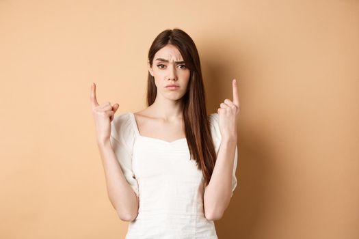Skeptical woman frowning and pointing fingers up, having doubts about product, standing on beige background. Copy space
