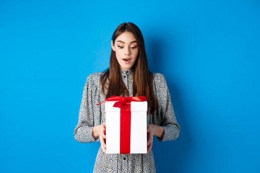Holidays. Surprised young woman looking at gift with amazed face, receive Valentines day present, standing in dress on blue background.
