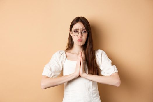 Sad girl in glasses begging for help, say please and looking cute, need favour, standing on beige background.