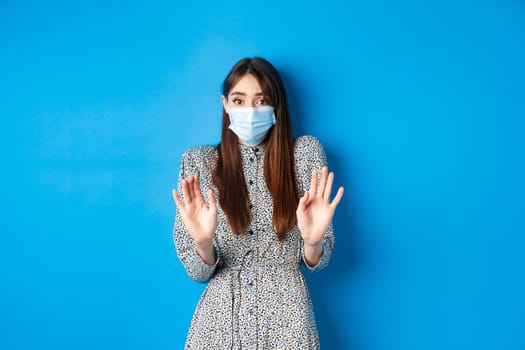 Covid-19, pandemic lifestyle concept. No thank you. Reluctant and worried girl in medical mask asking non-masker to stay away, showing block gesture, tell stop, blue background.