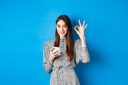 Beautiful young woman in dress showing okay sign and using smartphone, smiling at camera, standing on blue background.