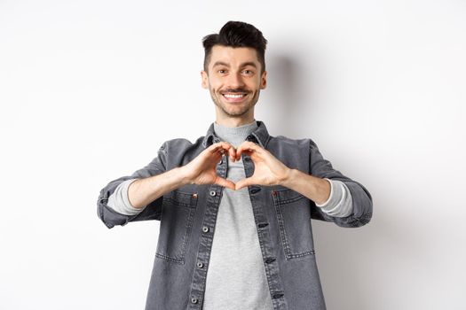Handsome man showing heart gesture and smiling. Romantic guy say I love you and smiling tender at camera. Valentines day concept.