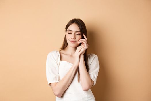 Tender and romantic woman close eyes, gently touching soft smooth skin, smiling dreamy while standing against beige background.
