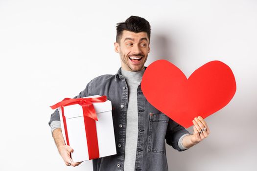 Happy boyfriend making romantic gifts on Valentines day date, holding box with present and big red heart card, smiling cheerful, white background.