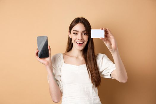 Cheerful young woman showing plastic credit card and empty mobile phone screen, smiling pleased at camera, standing on beige background.