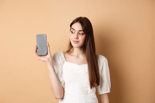 Attractive young woman showing empty smartphone screen, smiling and looking aside, standing on beige background.