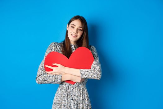 Valentines day. Dreamy romantic woman hugging big red heart cutout, looking sensual at camera, standing in dress on blue background.