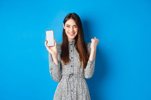 Cheerful pretty girl chanting, winning on mobile phone, showing empty smartphone screen and fist pump, smiling and celebrating, blue background.