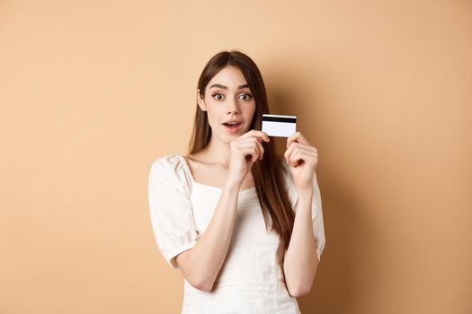 Excited young woman showing plastic credit card and look amazed at camera, standing on beige background.
