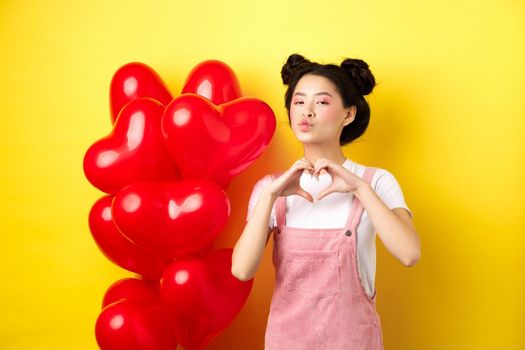 Beautiful asian woman showing heart, I love you gesture and kissing lips, standing near romantic red balloons. Concept of Valentines day.