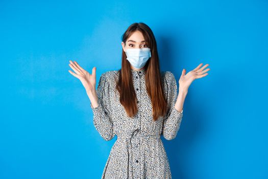 Healthy people and covid-19 pandemic concept. Surprised girl in medical mask raising hands up excited, hear good news, smiling with eyes, standing blue background.