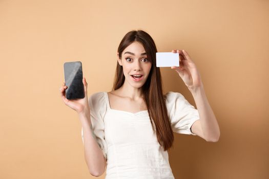 Excited girl showing plastic credit card of bank and empty mobile phone screen, demonstrate shopping app, standing on beige background.