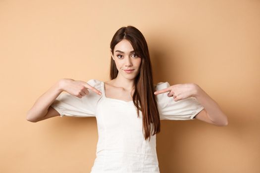 Young serious woman pointing at center, showing logo with confident face, standing in dress on beige background.