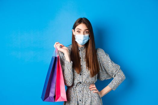 Covid-19, pandemic and lifestyle concept. Surprised woman wear medical mask on shopping, holding bags and look at camera, blue background.