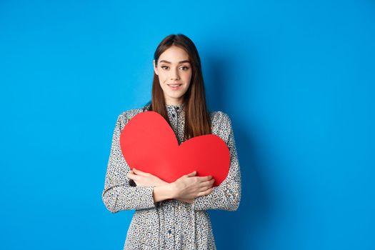 Valentines day. Attractive young woman searching for love, holding big red heart cutout and smiling at camera, standing in dress on blue background.