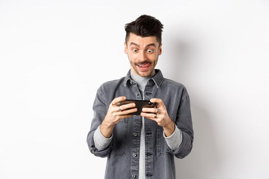 Excited guy playing video game on mobile phone with focused face, tap on smartphone screen, holding cellphone horizontally, white background.