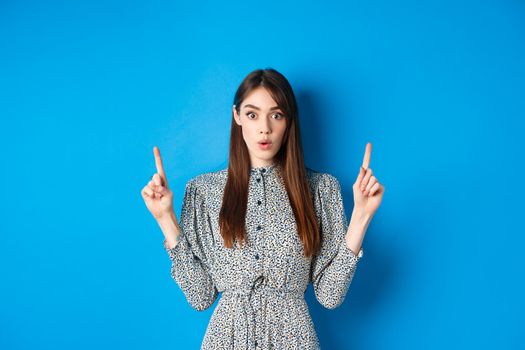 Excited pretty lady in dress say wow, looking intrigued by advertisement, pointing fingers up at logo, standing against blue background.
