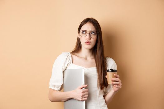 Confused and pensive girl with laptop looking aside suspicious, drinking takeaway coffee, standing against beige background.
