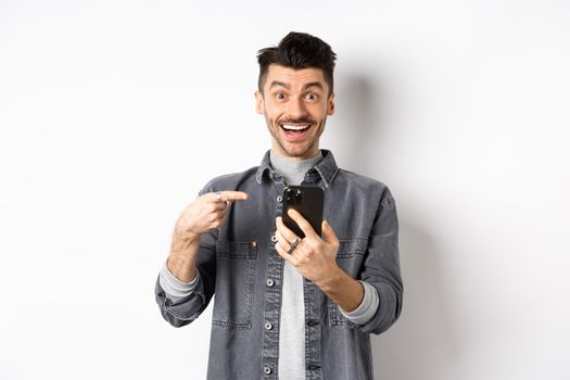 Excited young man receive good news on phone, pointing at smartphone and smiling happy, standing on white background.