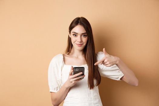 Attractive woman smiling and pointing at smartphone, showing online offer on phone, standing on beige background.